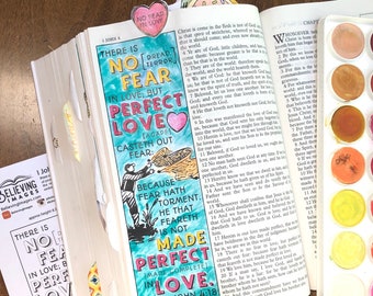 May include: An open Bible with a colorful bookmark that reads "NO FEAR IN LOVE." The page is hand-lettered with a quote about love casting out fear, and a watercolor palette is visible. The theme is faith and inspiration.