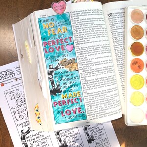 May include: An open Bible with a colorful bookmark that reads "NO FEAR IN LOVE." The page is hand-lettered with a quote about love casting out fear, and a watercolor palette is visible. The theme is faith and inspiration.