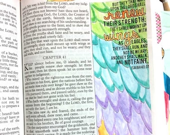 May include: Open Bible with a colorful, hand-painted bookmark featuring a wing design. The bookmark has text from Isaiah 40:31, with the words "renew their strength" and "mount up with wings as eagles."