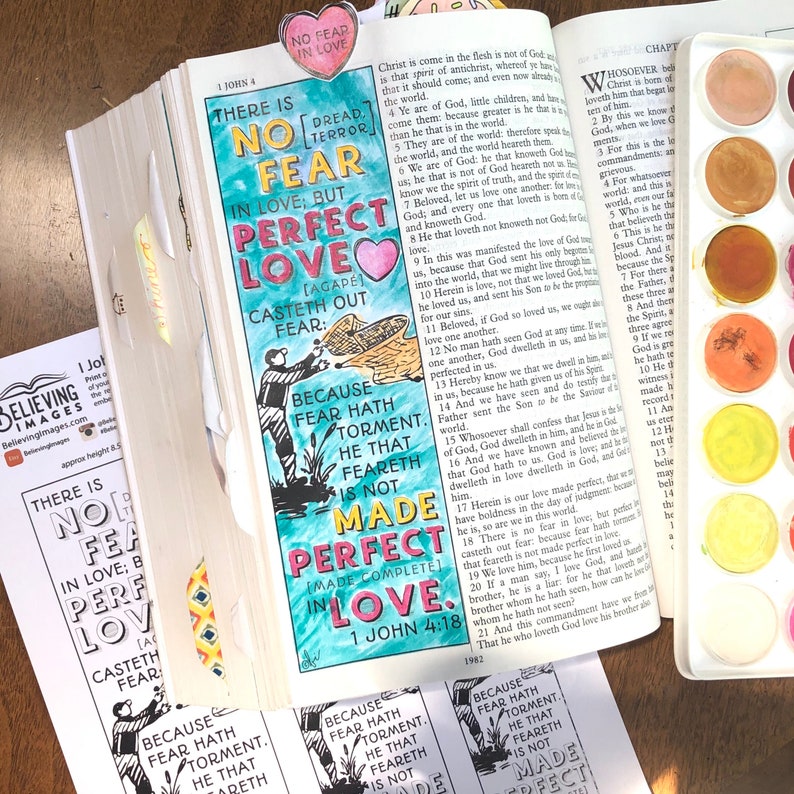 May include: An open Bible with a colorful bookmark that reads "NO FEAR IN LOVE." The page is hand-lettered with a quote about love casting out fear, and a watercolor palette is visible. The theme is faith and inspiration.
