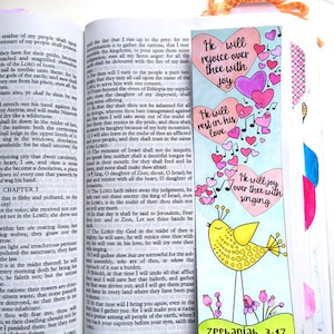 May include: A colorful bookmark with the text "He will rejoice over thee with joy," "He will rest in his love," and "He will joy over thee with singing." The bookmark features hearts, musical notes, and a yellow bird illustration. The text "Zephaniah 3:17" is also included.