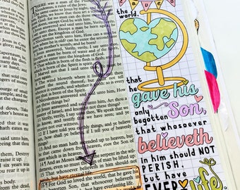 May include: Open Bible with colorful handwritten notes and illustrations. A globe illustration with the text "For God so loved the world" and "John 3:16". The page is marked with a purple ribbon and a heart-shaped paper cutout.