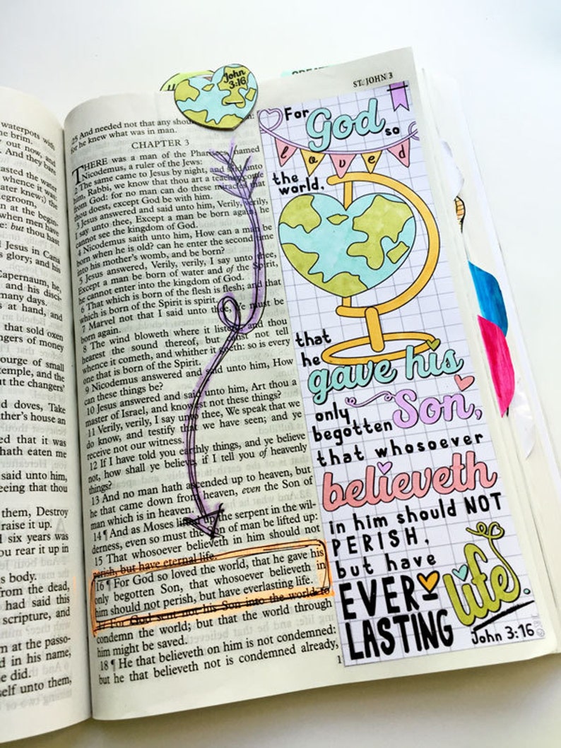 May include: Open Bible with colorful handwritten notes and illustrations. A globe illustration with the text "For God so loved the world" and "John 3:16". The page is marked with a purple ribbon and a heart-shaped paper cutout.
