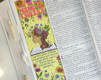 May include: A colorful floral bookmark with the text "PUT ON THE MIND OF CHRIST" and "PHILIPPIANS 2:5". The bookmark features a drawing of a woman with flowers in her hair, and is placed in an open book.