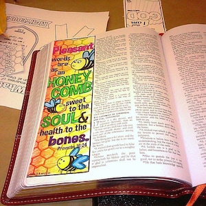 May include: A colorful bookmark with the text "Pleasant words are as an honeycomb sweet to the soul & health to the bones. Proverbs 16:24." The bookmark features cartoon bees and a honeycomb design, placed in an open Bible.