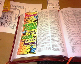 May include: A colorful bookmark with the text "Pleasant words are as an honeycomb sweet to the soul & health to the bones. Proverbs 16:24." The bookmark features cartoon bees and a honeycomb design, placed in an open Bible.