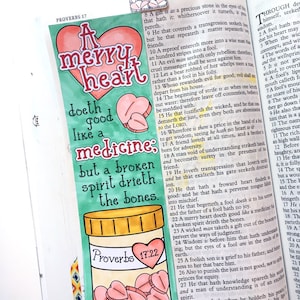 May include: A hand-drawn illustration of a heart with the words "A merry heart doth good like a medicine: but a broken spirit drieth the bones." The illustration is surrounded by a green border and a yellow pill bottle with pink hearts inside. The text "Proverbs 17:22" is written on the bottle.