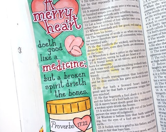 May include: A hand-drawn illustration of a heart with the words "A merry heart doth good like a medicine: but a broken spirit drieth the bones." The illustration is surrounded by a green border and a yellow pill bottle with pink hearts inside. The text "Proverbs 17:22" is written on the bottle.