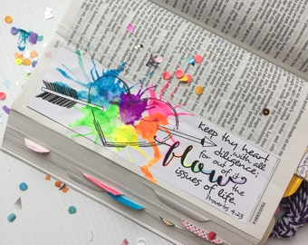 May include: A colorful bookmark with a heart and arrow design, featuring vibrant watercolor splashes. The text "keep thy heart with all diligence; for out of it are the issues of life" is displayed. The bookmark is placed in an open book.