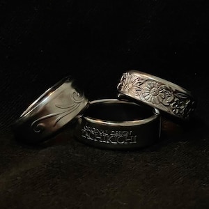 Puede incluir: Tres anillos de plata. Uno tiene un diseño floral, otro un patrón de remolino, y el tercero tiene grabado el texto "ORIGINAL DESIGN TACHIKICHI". Los anillos se muestran sobre un fondo oscuro.