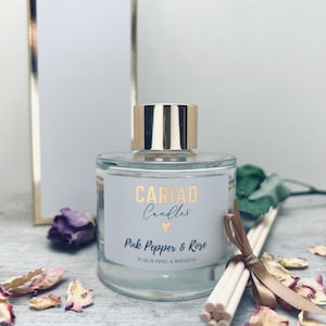 May include: A glass aromatherapy diffuser with a gold lid, labelled "CARIAD Candles Pink Pepper & Rose". The image shows dried rose petals and reeds tied with a ribbon, suggesting a floral scent.