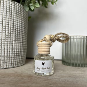 May include: A small, clear glass bottle with a wooden cap and a label that reads "Peony & Blush Suede" with a heart symbol. The bottle is suspended by a rope and wooden bead. The background includes a decorative vase and greenery.