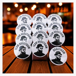Op de afbeelding: Een verzameling witte ronde badges met de tekst "TEAM BRÄUTIGAM" in een zwarte, vette letter. Elke badge heeft een zwart-witte illustratie van een man met een hoed en zonnebril. De badges liggen op een houten oppervlak.