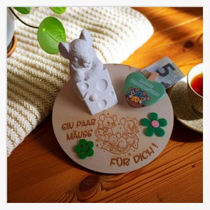 Op de afbeelding: Een houten decoratiestuk met muizen als thema, met een wit muizenbeeldje op een kaasblok. Het stuk bevat een groene bloem, een hartvormige magneet en de Duitse tekst "Ein Paar Mäuse für Dich!"