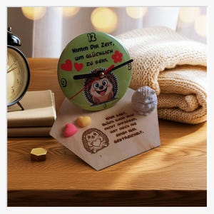 Op de afbeelding: Een decoratieve klok met een groene wijzerplaat met een cartoonegel en de Duitse tekst "Nimm Dir Zeit, um glücklich zu sein." De afbeelding bevat een houten plaat en hartvormige decoraties.