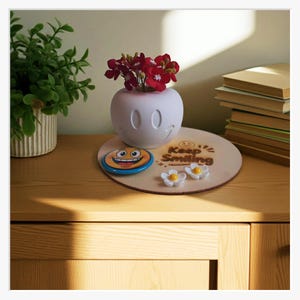 Könnte beinhalten: Eine weiße Vase mit Smiley-Gesicht und roten Blumen steht auf einem Holzuntersetzer mit dem Schriftzug "Keep Smiling". Ein blau-gelber Smiley-Untersetzer und zwei weiße Blumendekorationen sind ebenfalls vorhanden.