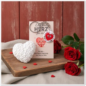 May include: A white textured heart-shaped object sits next to a keychain with a round charm featuring a red cartoon heart. A card behind the keychain displays the text "Ein kleines HERZ für dich" and "Für dich - eine kleine Umarmung von Herz zu Herz."
