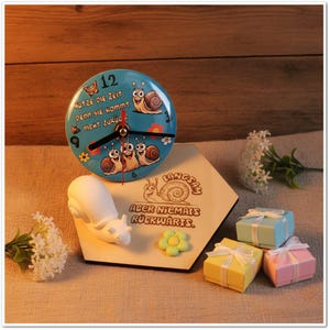 Puede incluir: Un reloj caprichoso con una esfera azul claro con caracoles de dibujos animados y el texto "NUTZE DIE ZEIT, DENN SIE KOMMT NICHT ZURÜG!". Se encuentra sobre una base de madera con un diseño de caracol y las palabras "LANGSAM ABER NIEMALS RÜCKWÄRTS."