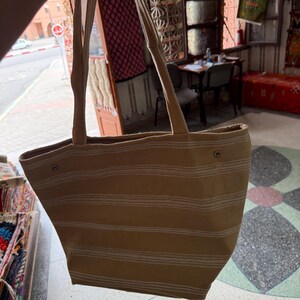 May include: A beige tote bag with horizontal white stripes. The bag has long, beige straps and is held up by a hand. The background shows a shop with rugs and a street scene.