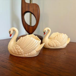 Puede incluir: Dos figuras de cisnes de color marfil con picos rojos. Los cisnes tienen diseños de plumas detallados y están colocados sobre una superficie de madera. El fondo incluye una lámpara de madera y una pared blanca. Estos cisnes decorativos son probablemente vintage o retro.
