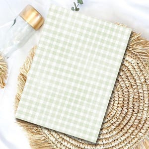 May include: A rectangular notebook featuring a light sage green and white gingham check pattern. The notebook is positioned on a round, woven straw placemat with a fringed edge, accompanied by a clear glass bottle with a metallic gold cap.