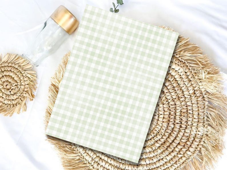 May include: A rectangular notebook featuring a light sage green and white gingham check pattern. The notebook is positioned on a round, woven straw placemat with a fringed edge, accompanied by a clear glass bottle with a metallic gold cap.
