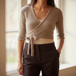 May include: A light brown wrap sweater with a tie closure, paired with dark brown trousers. The sweater has three-quarter sleeves and a V-neck design. The model is standing near a window.