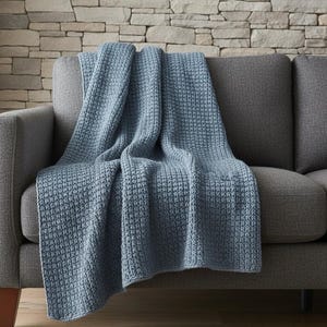 May include: A blue woven throw blanket draped over a gray sofa. The blanket has a textured, waffle-like pattern. The sofa has a modern design with a low profile and wooden legs. The background features a stone wall.