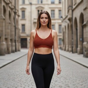 May include: A woman wearing a rust-colored knitted crop top and black leggings walks down a cobblestone street. The buildings on either side are made of stone and have arched doorways.