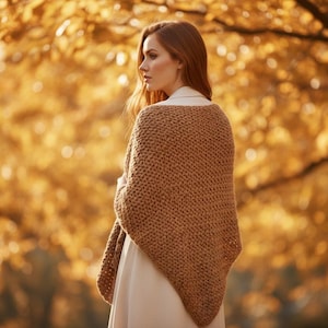 May include: A woman wearing a brown knitted shawl over a white dress, standing outdoors. The shawl is a solid color with a chunky knit texture. The background features blurred golden-yellow foliage.