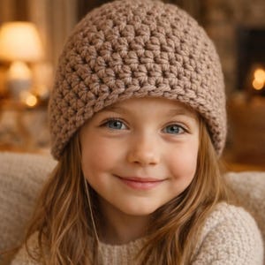 May include: A young person wearing a tan crochet beanie. The beanie is made of a thick, textured yarn and fits snugly on the head. The person has long, light brown hair and is wearing a light-colored sweater. The background is softly blurred with warm lighting.