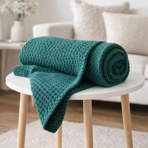 May include: A rolled-up teal blanket with a textured pattern rests on a small, white, round table with wooden legs. The blanket is partially unrolled, displaying its soft, woven texture. The background includes a white sofa and a vase of flowers.
