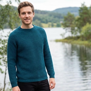 May include: A teal ribbed knit sweater with a crew neck. The long-sleeved sweater is worn by a person standing outdoors near a lake. The sweater is a solid color and has a classic, casual style.
