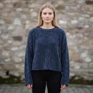 May include: A woman wearing a navy blue, ribbed-knit sweater with a crew neck and long sleeves. The sweater is paired with black pants. The background is a stone wall.