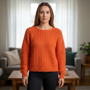 May include: An orange, chunky knit sweater with a crew neck. The sweater has a ribbed texture and long sleeves. The person is wearing black pants. The background includes a couch, a table, and a lamp.