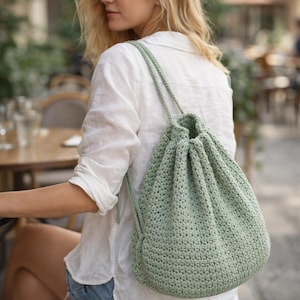 May include: A light green crochet drawstring backpack with shoulder straps. The bag is made of a textured, open-weave stitch and has a rounded shape. The drawstring closure is visible at the top. The bag is worn over the shoulder.