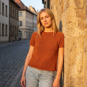 May include: A short-sleeved, rust-colored knit top. The top has a boat neck and a ribbed texture. The person is wearing light blue jeans and is leaning against a stone wall in an outdoor setting. The background shows a cobblestone street and buildings.