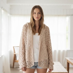May include: A woman wearing a beige, open-weave hooded cardigan over a white top and denim shorts. The cardigan has a loose, relaxed fit. The background features a bright, airy room with large windows and white curtains.