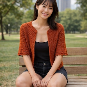 Puede incluir: Un cárdigan de ganchillo de color óxido con mangas cortas. El suéter abierto tiene un patrón de puntadas texturizadas y un borde festoneado en las mangas. La persona lleva una camiseta sin mangas negra y pantalones cortos de mezclilla, sentada en un banco de madera.