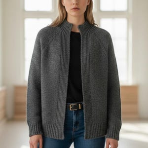 May include: A gray, open-front cardigan with a ribbed texture. The cardigan has a high, rounded collar and long sleeves. The model is wearing a black shirt and blue jeans. A black belt with a gold buckle is visible.