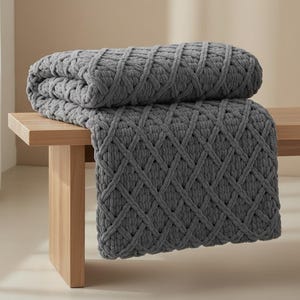 May include: A folded gray knitted blanket with a diamond pattern rests on a light wood bench. The blanket's thick yarn creates a textured, cozy appearance. The bench has a simple, modern design.