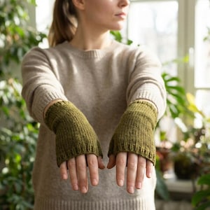 May include: Olive green knitted fingerless gloves. The gloves are made of a textured yarn and have a ribbed cuff. The person is wearing a light brown sweater. The background is filled with green plants.