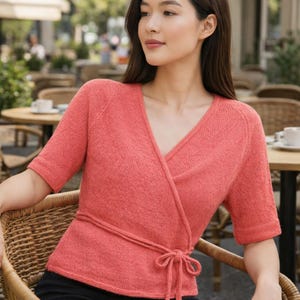 May include: A coral-colored wrap top with short sleeves and a V-neck. The top features a tie closure at the waist and is made of a textured knit fabric. The model is seated in a wicker chair, suggesting a casual, everyday style.