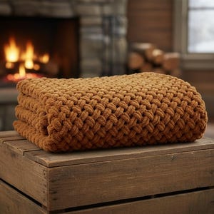 May include: A folded, chunky knit blanket in a warm, earthy brown hue. The blanket is made of thick yarn, creating a textured, woven appearance. It rests on a rustic wooden crate, with a fireplace in the background.