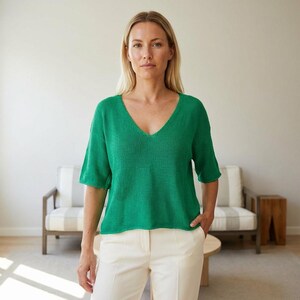 May include: A woman wearing a short-sleeved, emerald green V-neck sweater and white pants. The sweater has a textured knit and a relaxed fit. The woman is standing in front of a neutral-toned background.