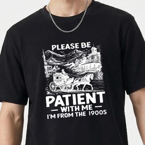 Puede incluir: Camiseta negra con un gráfico blanco que presenta un carruaje tirado por caballos y el texto "PLEASE BE PATIENT WITH ME - I'M FROM THE 1900S". El diseño incluye una ilustración vintage de una ciudad y montañas.