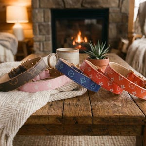 May include: A collection of dog collars in various colors, including brown, pink, blue, and orange. Each collar features a repeating pattern and gold-colored hardware. The collars are displayed on a wooden table with a cozy, rustic aesthetic.