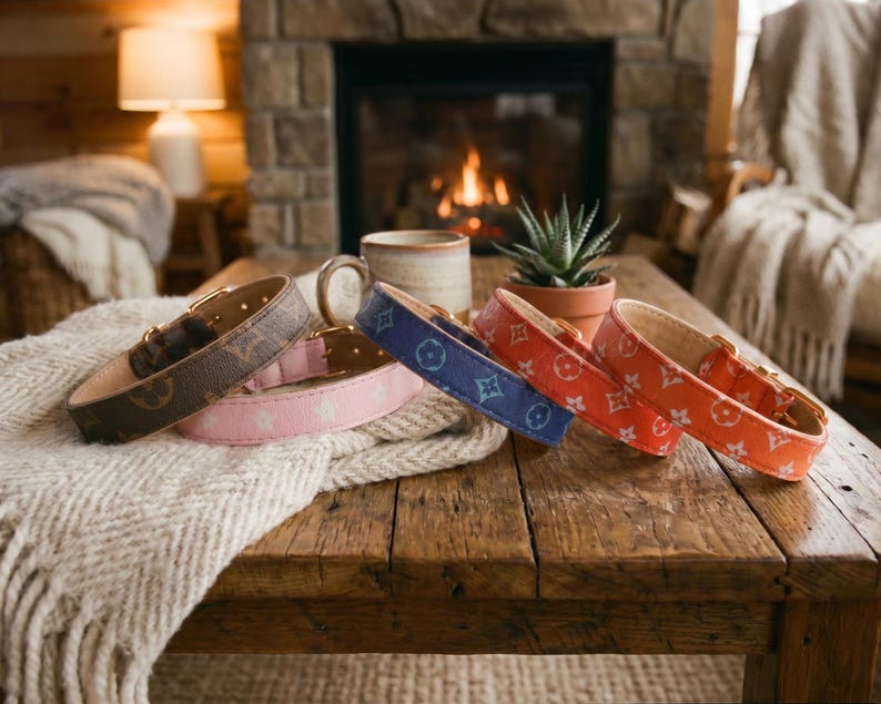 May include: A collection of dog collars in various colors, including brown, pink, blue, and orange. Each collar features a repeating pattern and gold-colored hardware. The collars are displayed on a wooden table with a cozy, rustic aesthetic.