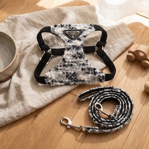 May include: A dog harness and lead set in a black and white tweed-style fabric. The harness has black straps and metal fixings. The lead is coiled, with a silver clasp. A ceramic bowl and wooden toys are also in the image.