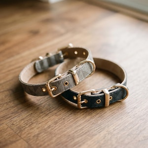 May include: Two pet collars featuring a repeating geometric pattern. One collar is light gray and the other is dark charcoal gray. Both collars include gold-toned metal buckles, D-rings, and eyelets, with a light-colored interior lining visible.